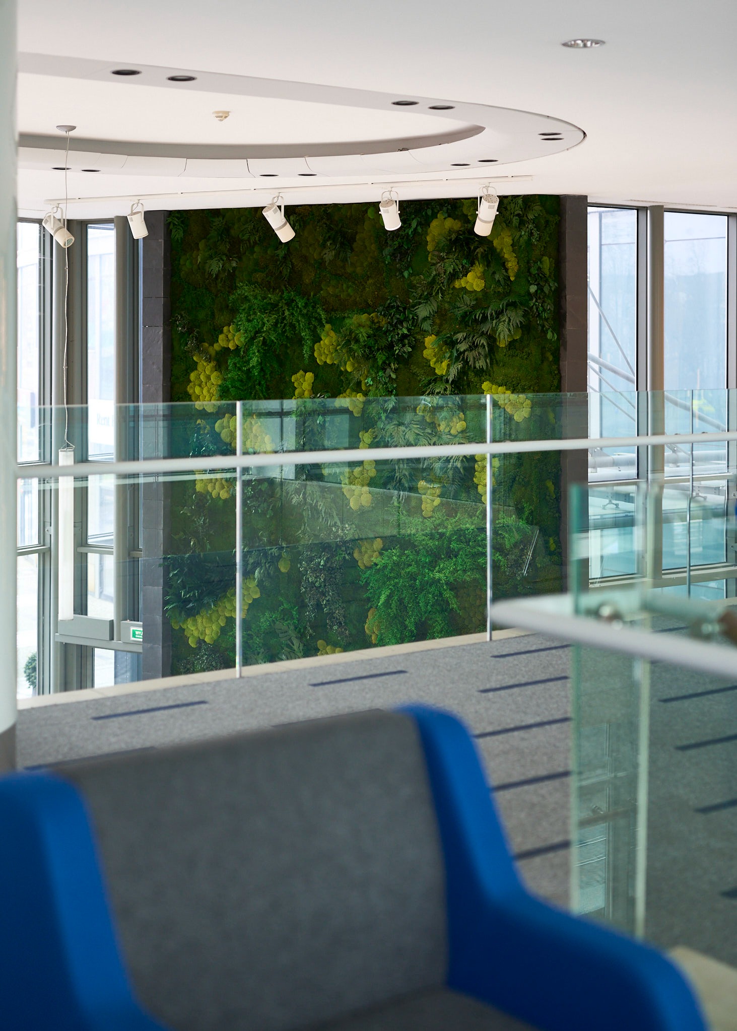 Wnętrze biura z zieloną ścianą z mchu, oświetloną reflektorami. Widok z góry przez szklaną balustradę, w tle okna i fragment sofy.