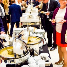 u.studio Paulina Miziewicz - Bufet z podgrzewaczami i zupą na firmowym evencie. Osoby w tle wybierają jedzenie. Stosy białych filiżanek i talerzy czekają na gości. Elegancki catering na spotkaniu biznesowym.