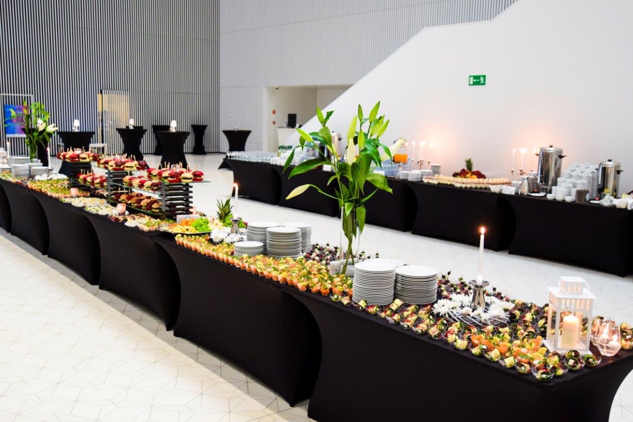 Długi stół cateringowy z czarnym obrusem, bogato zastawiony przekąskami, kanapkami i dekoracjami kwiatowymi w nowoczesnym wnętrzu z białymi ścianami i geometryczną podłogą.