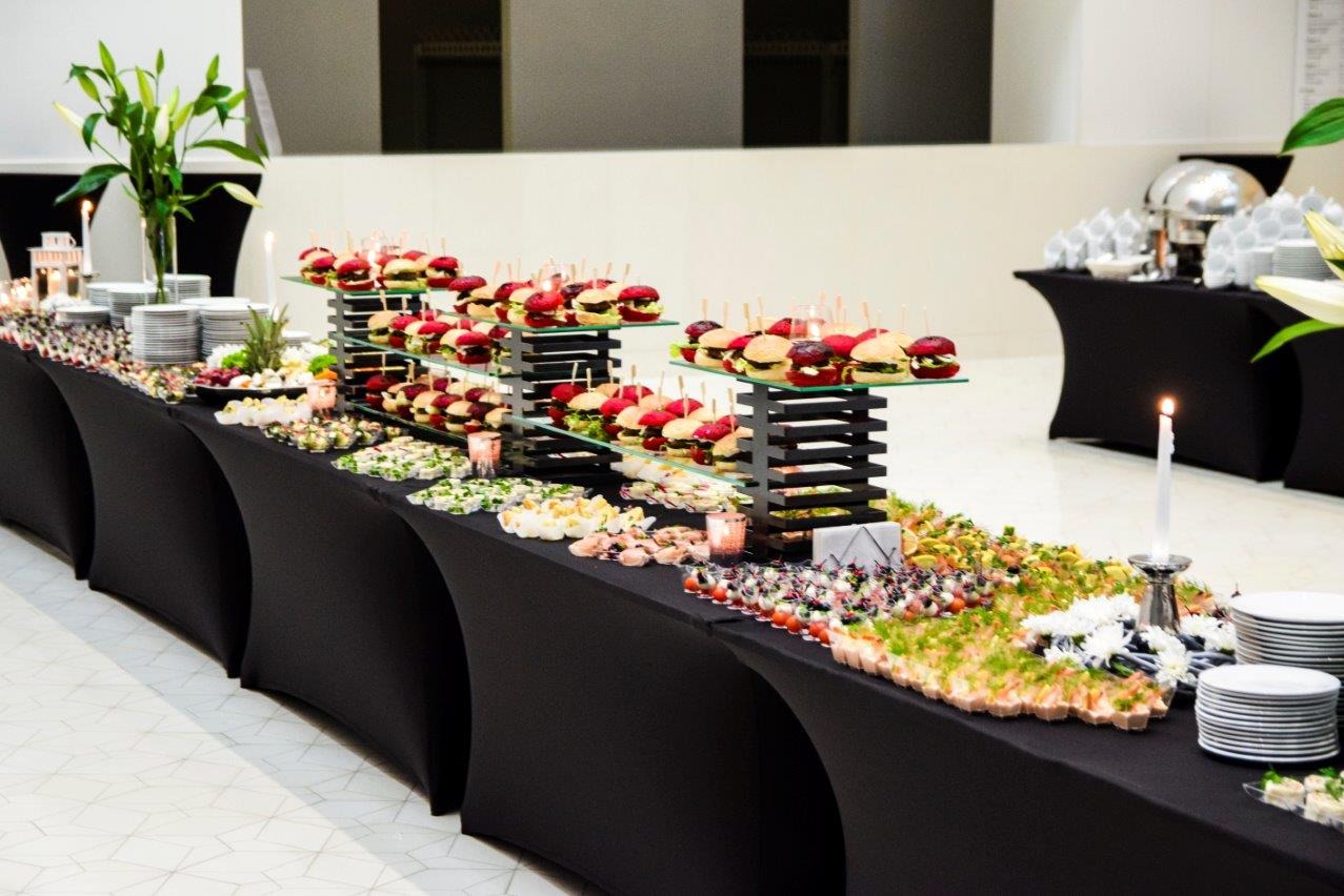 Elegancki stół cateringowy z mini burgerami, sałatkami i przekąskami na czarnym obrusie, oświetlony świecami, tworzący wyrafinowaną aranżację.