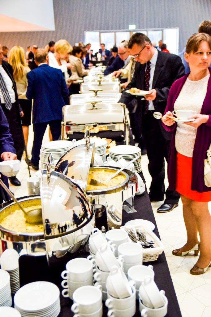 Bufet z podgrzewaczami i zupą na firmowym evencie. Osoby w tle wybierają jedzenie. Stosy białych filiżanek i talerzy czekają na gości. Elegancki catering na spotkaniu biznesowym.