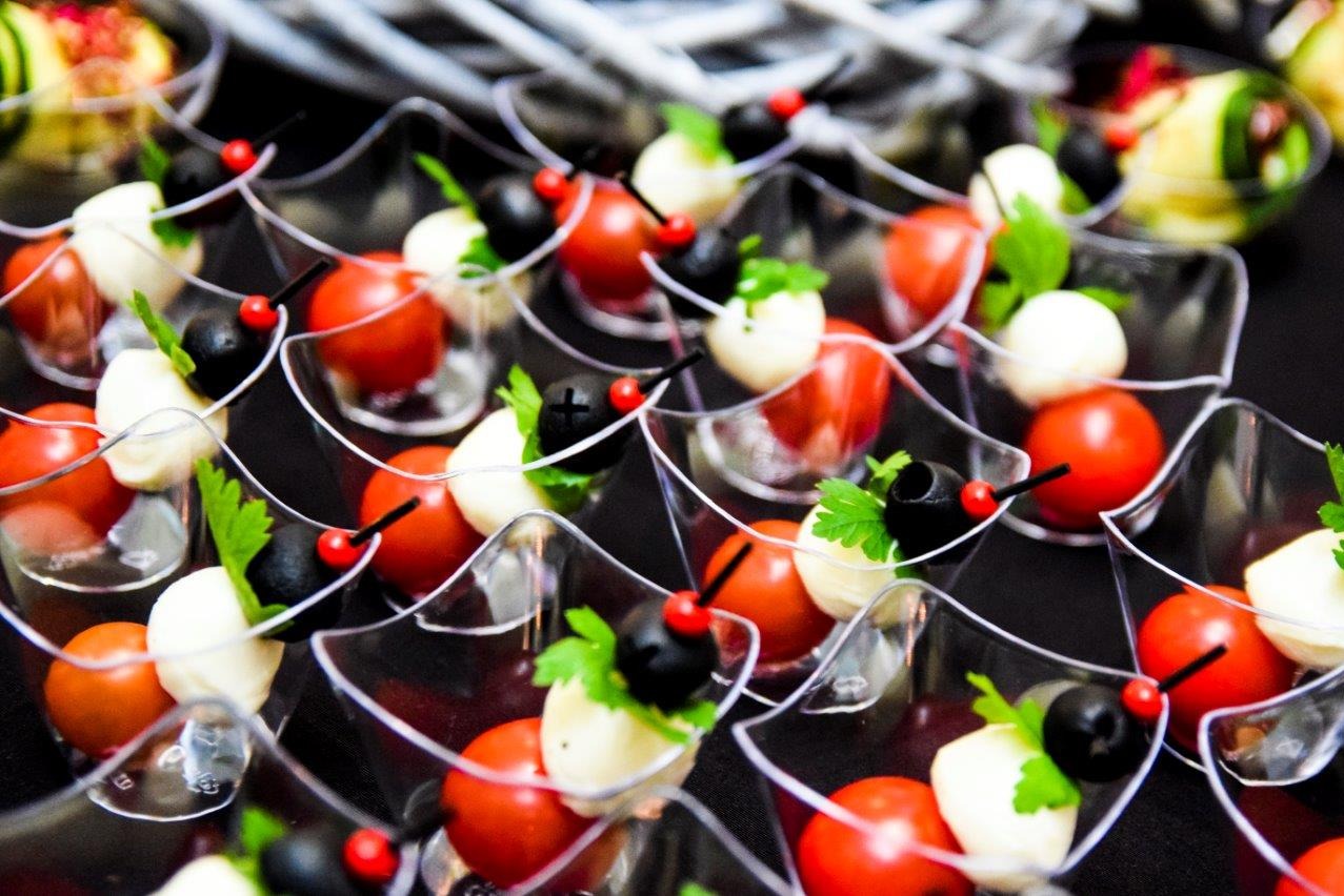 Miniaturowe przekąski caprese w przezroczystych naczyniach: pomidorki cherry, mozzarella, czarne oliwki i listki bazylii, elegancko podane na ciemnym tle.