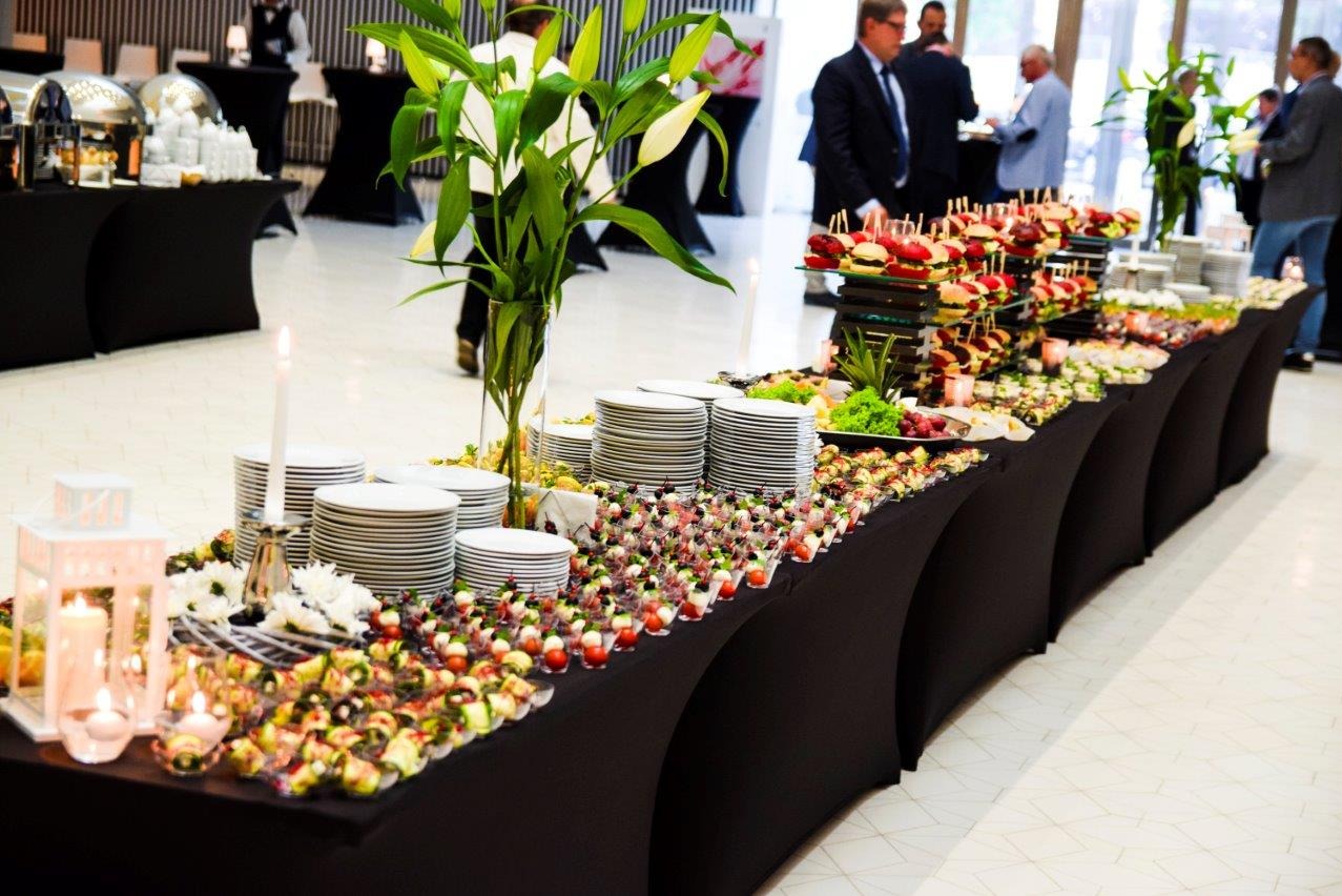 Elegancko zastawiony stół cateringowy z przekąskami, dekoracjami kwiatowymi i świecami, gotowy na przyjęcie. Widoczni goście w tle.