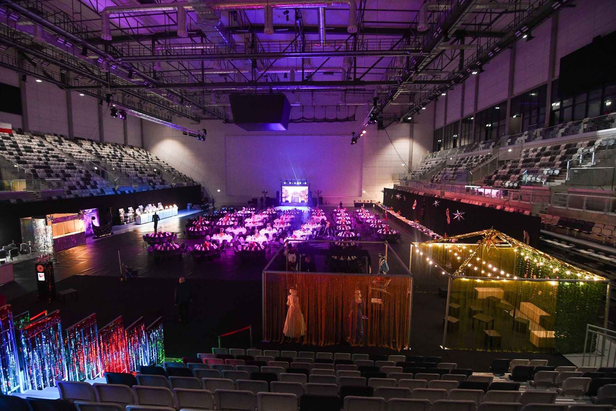 Widok z góry na halę widowiskową przygotowaną na galę. Ustawione okrągłe stoły, dekoracje świetlne i ścianki działowe. Scena w tle. Fioletowe oświetlenie.