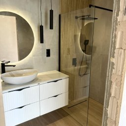 New Vision Pro Artur Froncz - Nowoczesna łazienka z drewnianymi akcentami: kabina prysznicowa, okrągłe lustro z podświetleniem LED i biała szafka pod umywalką. Widoczny fragment ściany w stanie surowym.