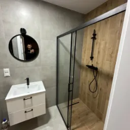 Nowoczesna łazienka z kabiną prysznicową typu walk-in z czarnymi profilami, umywalką z szafką i okrągłym lustrem z odbiciem osoby.