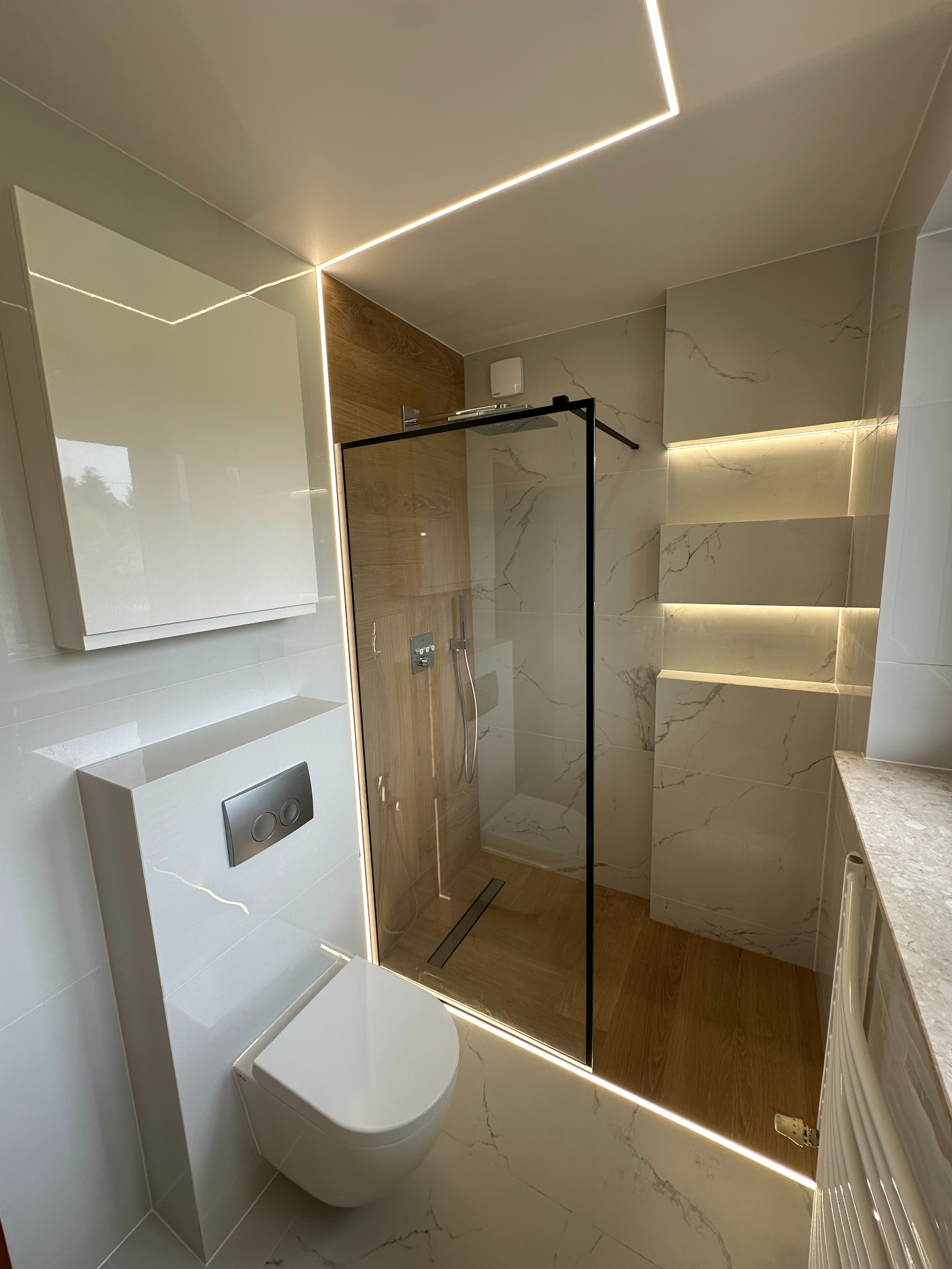 Nowoczesna łazienka z kabiną prysznicową typu walk-in, podwieszaną toaletą i oświetleniem LED wzdłuż sufitu i wnęk. Marmurowe płytki i drewniane akcenty.