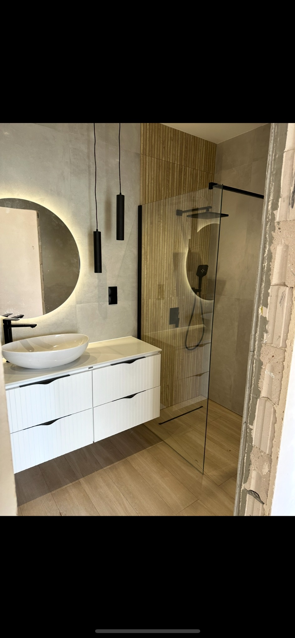Nowoczesna łazienka z drewnianymi akcentami: kabina prysznicowa, okrągłe lustro z podświetleniem LED i biała szafka pod umywalką. Widoczny fragment ściany w stanie surowym.