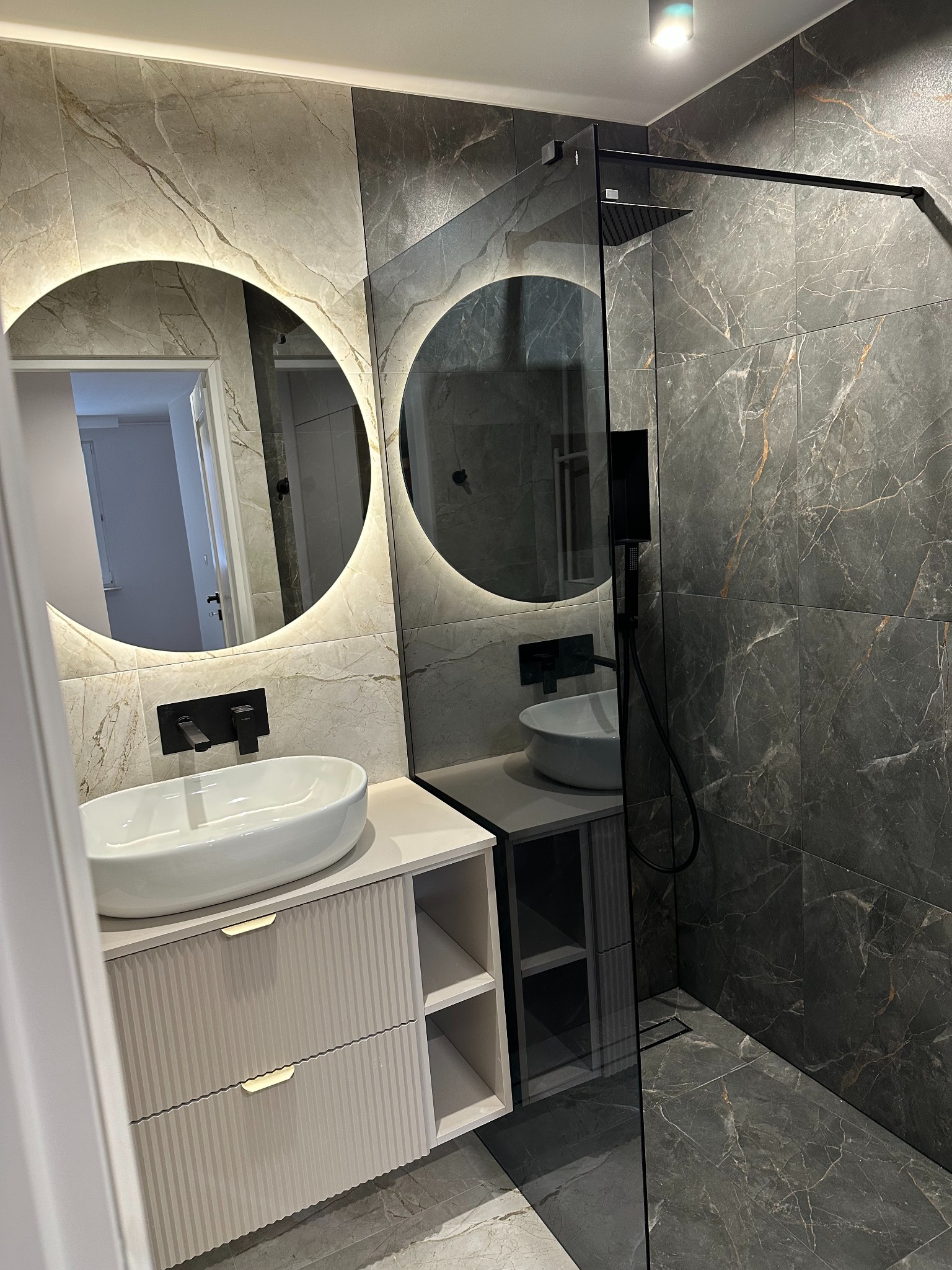 Nowoczesna łazienka z marmurowymi płytkami, okrągłym lustrem z podświetleniem LED, białą umywalką nablatową i czarną armaturą, kabina prysznicowa typu walk-in.