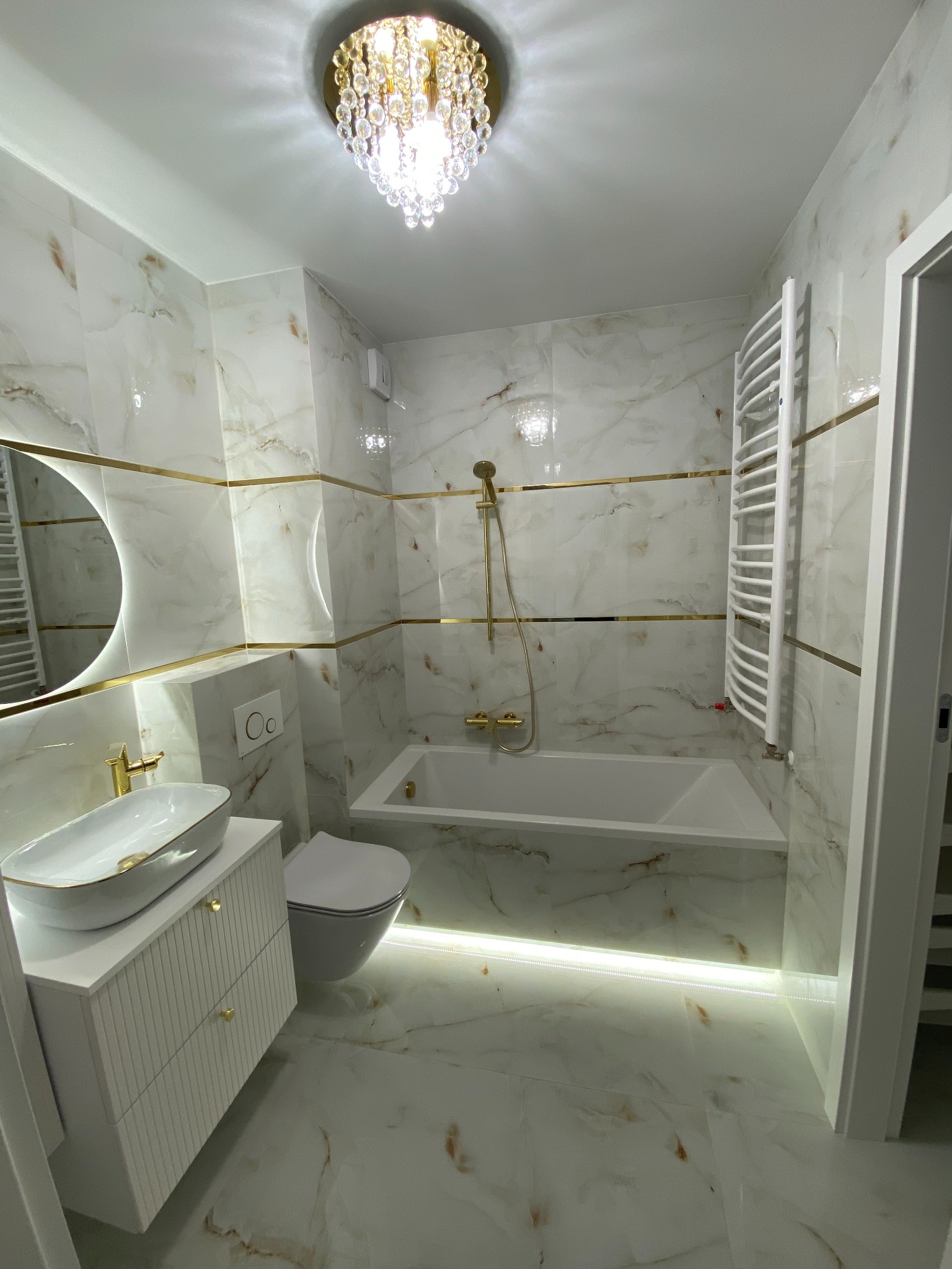 Elegancka łazienka w bieli i złocie: marmurowe płytki, podświetlana podłoga LED, okrągłe lustro i kryształowy żyrandol. Nowoczesny design z luksusowymi akcentami.