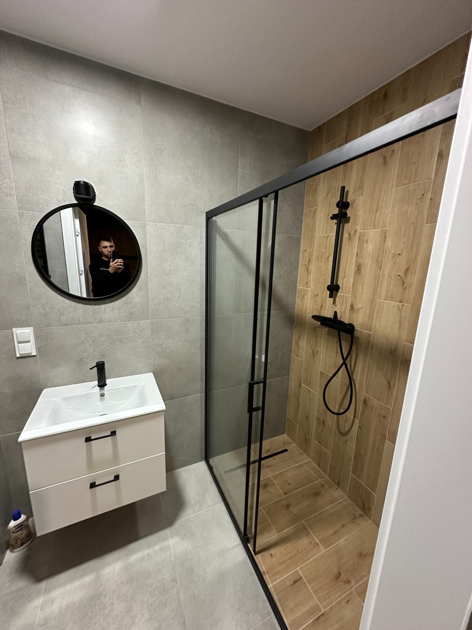 Nowoczesna łazienka z kabiną prysznicową typu walk-in z czarnymi profilami, umywalką z szafką i okrągłym lustrem z odbiciem osoby.