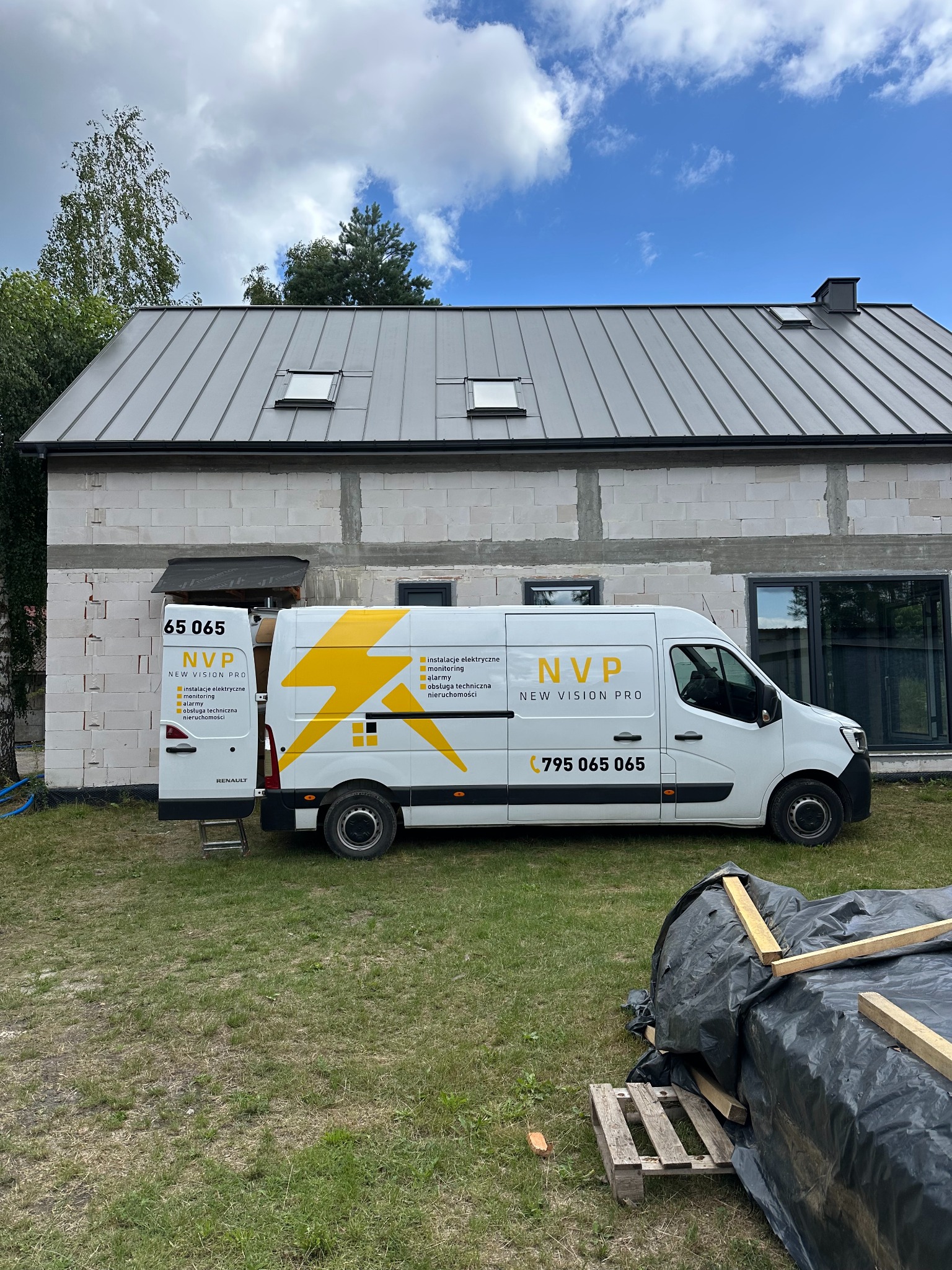 Biały van z logo NVP New Vision Pro na tle surowego domu z białej cegły. Otwarty tył vana z listą usług: instalacje elektryczne, monitoring, alarmy.