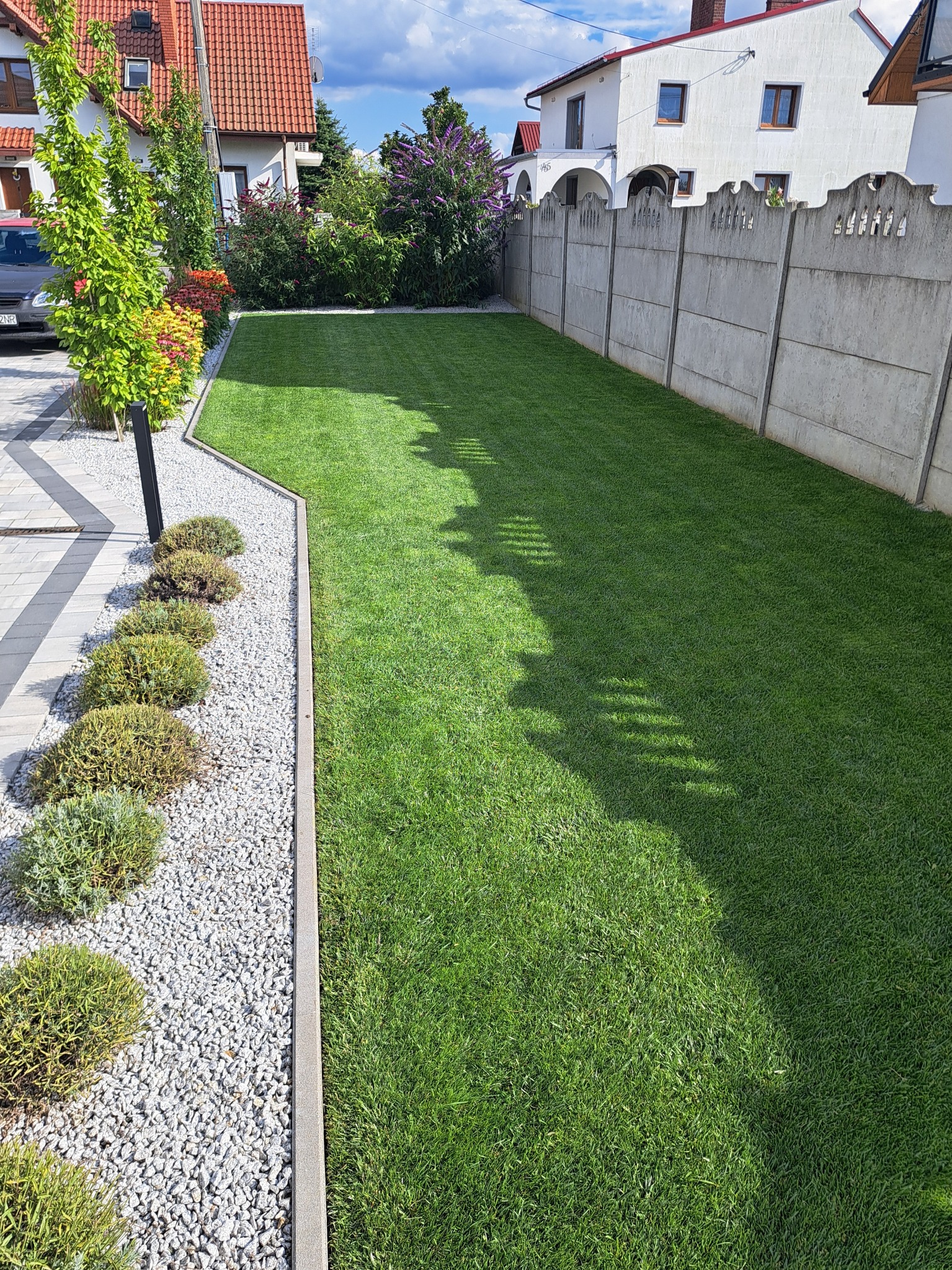 Perfekcyjnie utrzymany trawnik z kosiarką wrzecionową, rabata z nasadzeniami i krawędziowaniem, oddzielony od ścieżki żwirowej betonowym obrzeżem.