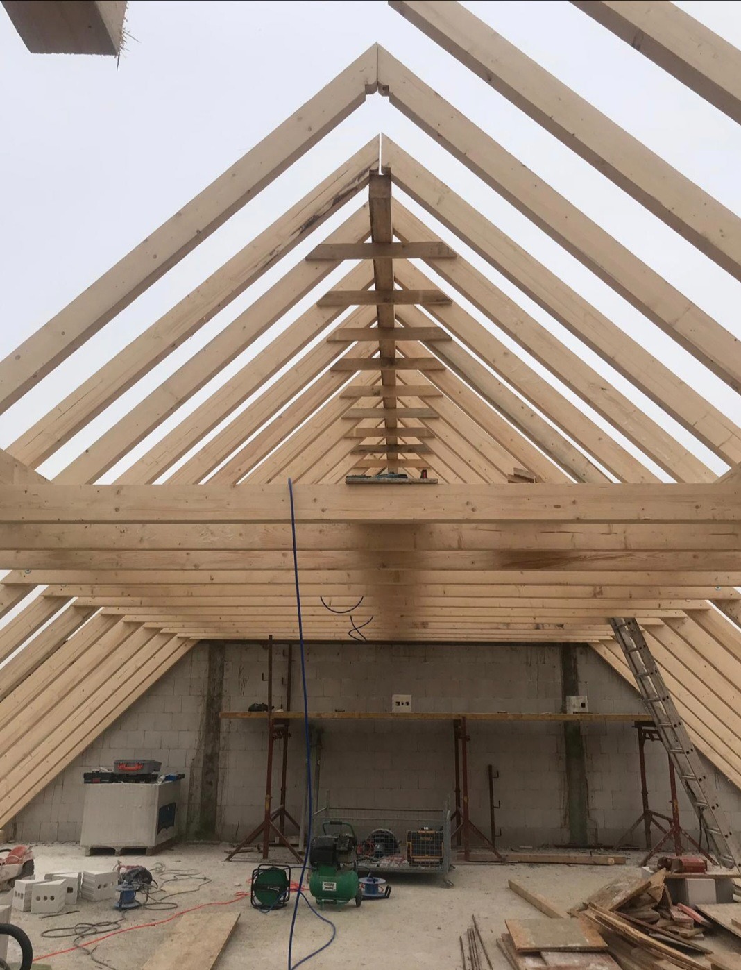 Drewniana konstrukcja dachu w trakcie budowy, widok od wewnątrz. Jasne drewno belek na tle surowych ścian i sprzętu budowlanego.