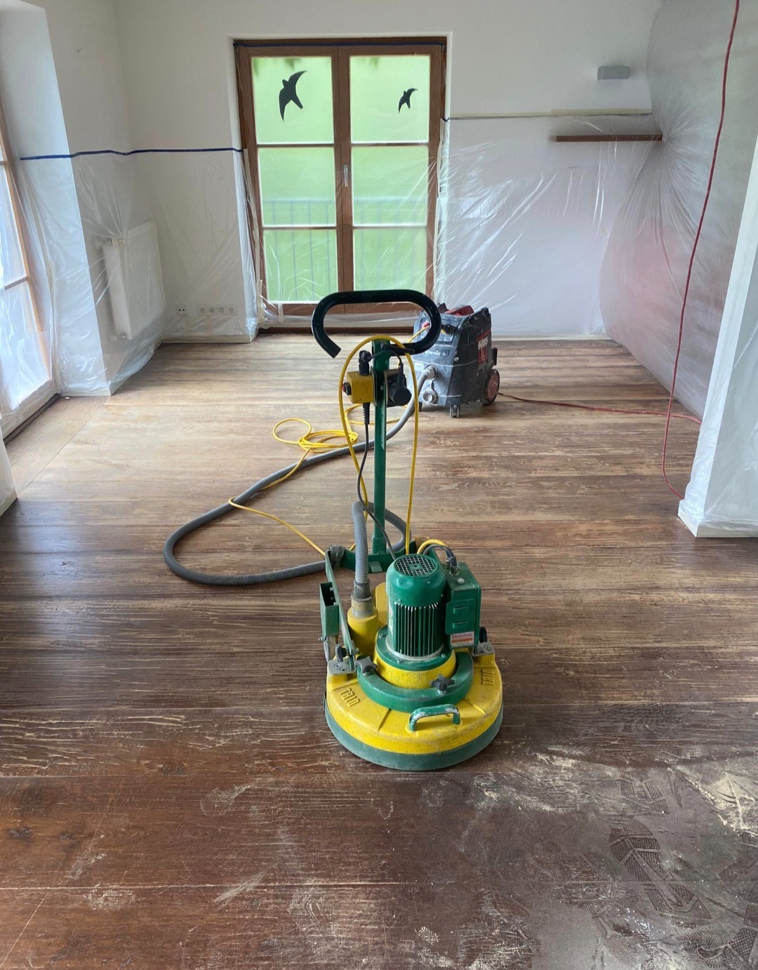 Szlifowanie drewnianej podłogi w pokoju zabezpieczonym folią malarską. Widoczna szlifierka parkieciarska i odkurzacz przemysłowy. Na oknie naklejki z ptakami.
