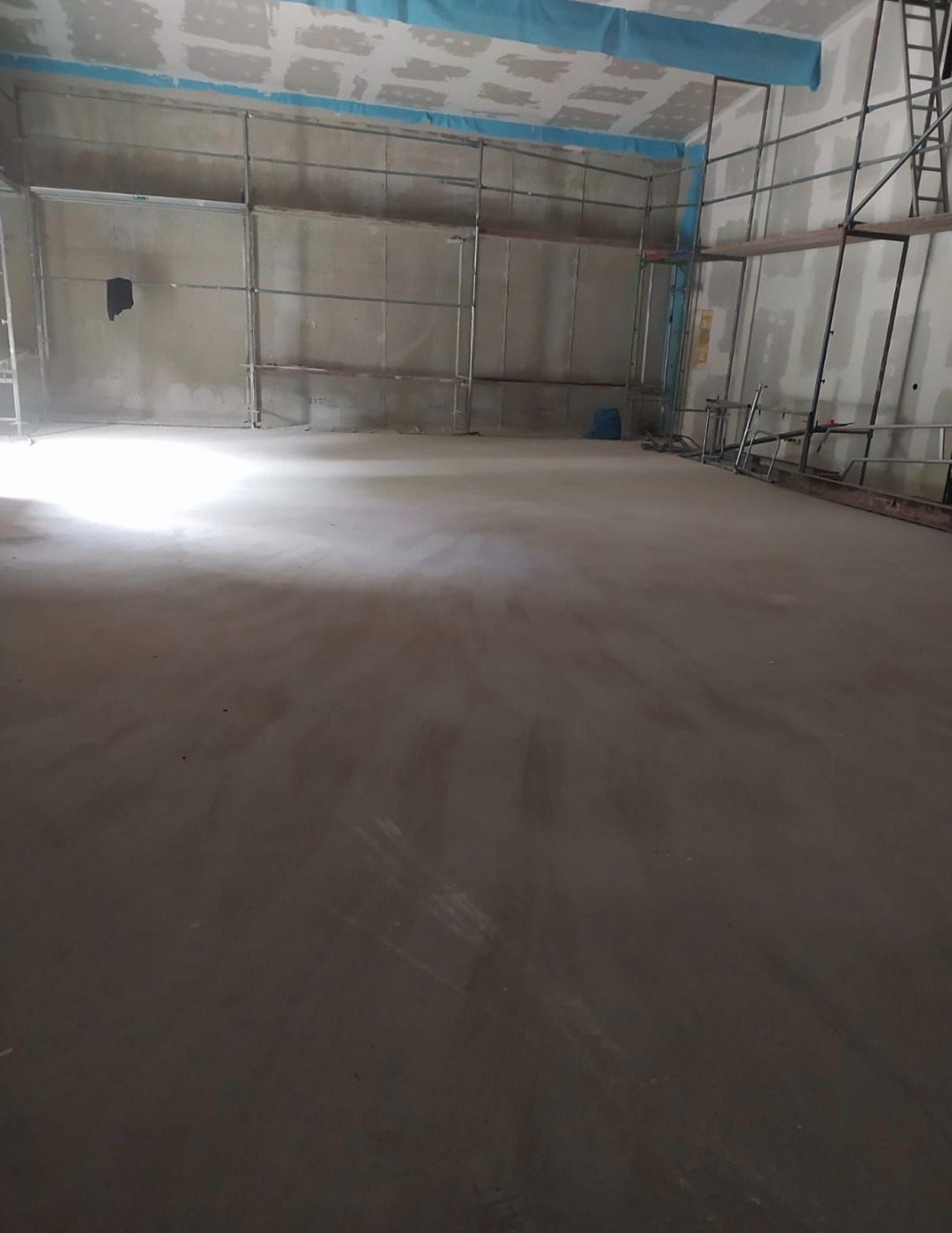 Wnętrze w trakcie szpachlowania z rusztowaniami. Sufit zabezpieczony folią. Podłoga wylewka. Ściany przygotowane pod malowanie. Widok na dużą powierzchnię w trakcie prac wykończeniowych.