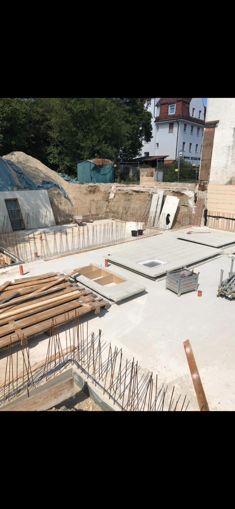 Fundamenty domu w trakcie budowy: zbrojenie, szalunki, beton. Na dalszym planie sterta gruzu i budynek mieszkalny. Dzień słoneczny.