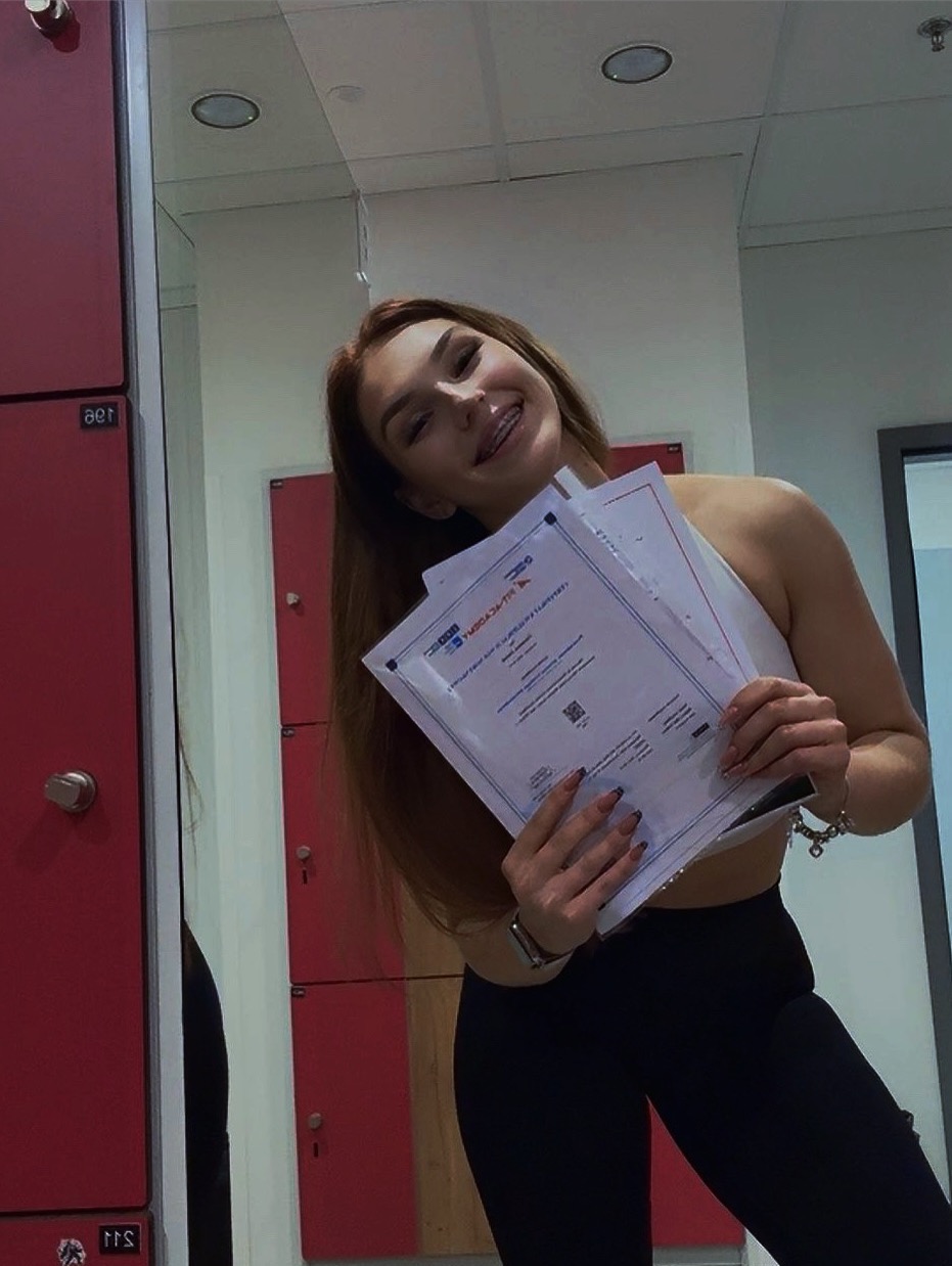 Uśmiechnięta kobieta z aparatem ortodontycznym, trzymająca certyfikaty w szatni siłowni, ubrana w sportowy top i legginsy. Widoczne czerwone szafki.