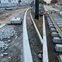 TELECOMSLOWI & REALESTETE Sp. z o.o. - Fragment budowy torowiska tramwajowego z ułożonymi obrzeżami z kostki granitowej i przygotowaną podbudową. W tle budynki mieszkalne i sprzęt budowlany.