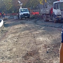 TELECOMSLOWI & REALESTETE Sp. z o.o. - Przygotowanie terenu pod budowę: wyrównana, kamienista powierzchnia z ciężarówkami i podnośnikiem koszowym w tle. Widoczni pracownicy w odblaskowych kamizelkach.