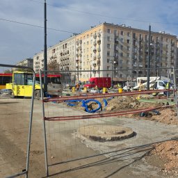 TELECOMSLOWI & REALESTETE Sp. z o.o. - Teren budowy z wykopem, widoczne niebieskie rury, maszyny budowlane i budynek mieszkalny w tle, zabezpieczony metalowym ogrodzeniem tymczasowym.