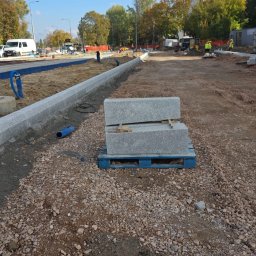 TELECOMSLOWI & REALESTETE Sp. z o.o. - Układanie obrzeży chodnika z kostki granitowej na placu budowy. Ułożone bloki na palecie, w tle ekipa pracowników i sprzęt budowlany, błękitne niebo.