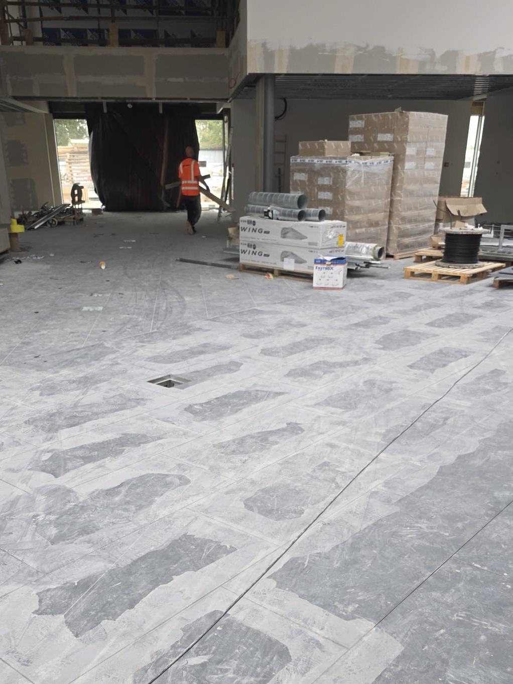 Wnętrze w trakcie wykańczania: szara podłoga z wyznaczonymi polami, materiały budowlane na paletach, pracownik w oddali. Widoczny surowy stan budynku.