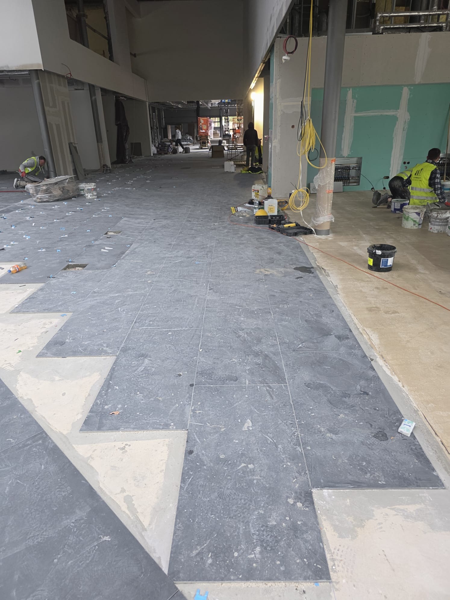 Układanie ciemnych płytek podłogowych w długim korytarzu. Widoczne narzędzia i materiały budowlane. Dwóch pracowników w trakcie pracy.