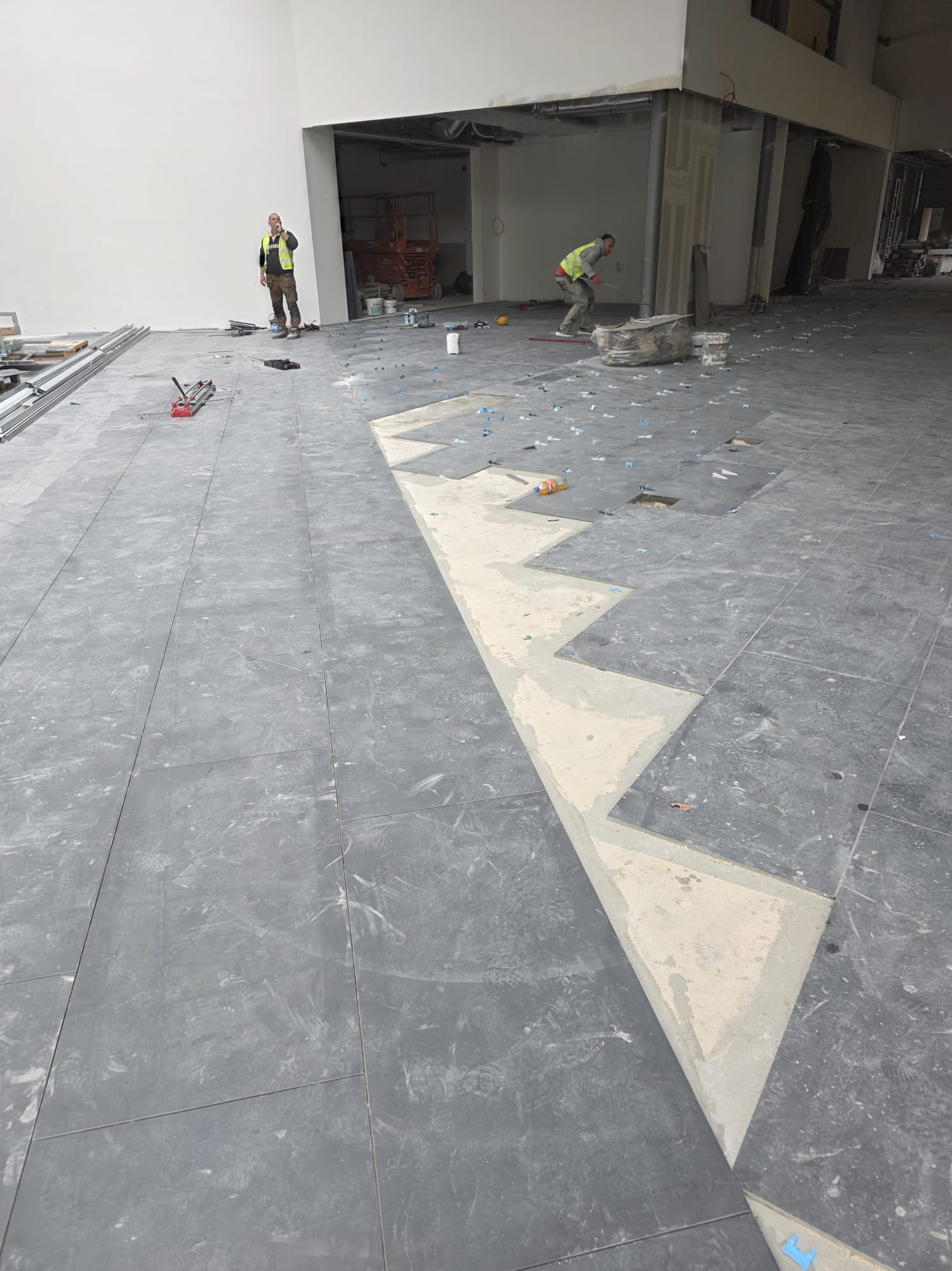 Układanie szarej, dużej płytki podłogowej w przestronnym wnętrzu. Widoczni dwaj pracownicy w odblaskowych kamizelkach i narzędzia glazurnicze.