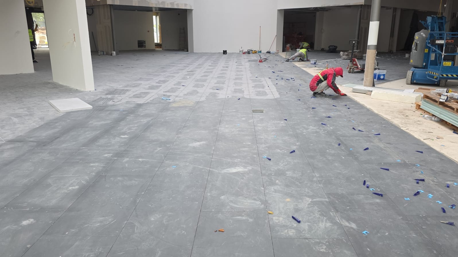 Układanie ciemnych płytek podłogowych w przestronnym wnętrzu. Pracownik w czerwonym kombinezonie układa płytki. Widoczne kliny dystansowe i narzędzia glazurnicze.