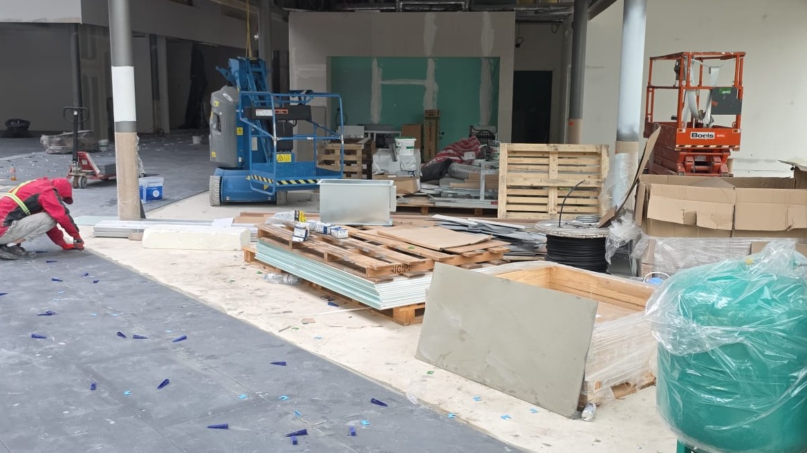 Pracownik w czerwonej kurtce układa ciemne płyty, używając niebieskich klipsów dystansowych. W tle widoczne materiały budowlane i podnośniki.