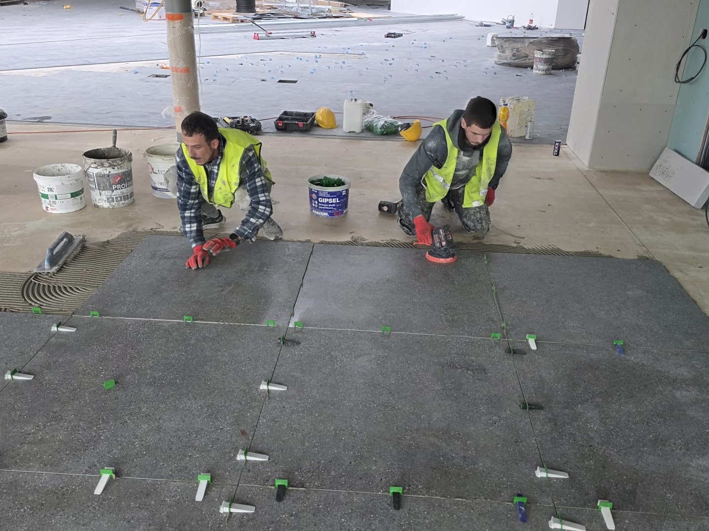 Dwóch glazurników w odblaskowych kamizelkach układa szare płytki podłogowe, używając poziomujących klipsów i narzędzi do dociskania. Widoczne wiadra z zaprawą i narzędzia.