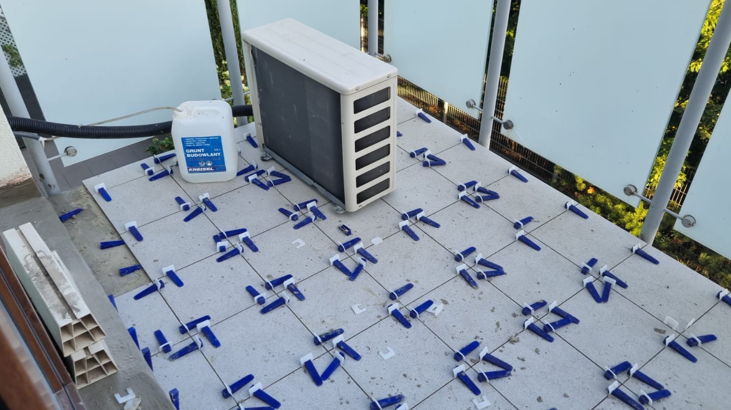 Balkon w trakcie kładzenia płytek. Widoczne krzyżyki glazurnicze, grunt budowlany Kreisel i jednostka zewnętrzna klimatyzacji. Jasne płytki i szklana balustrada.