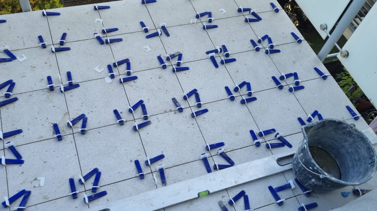 Jasne płytki ułożone z użyciem niebieskich klinów poziomujących, wiaderko i poziomica w kadrze. Etap prac glazurniczych na balkonie.