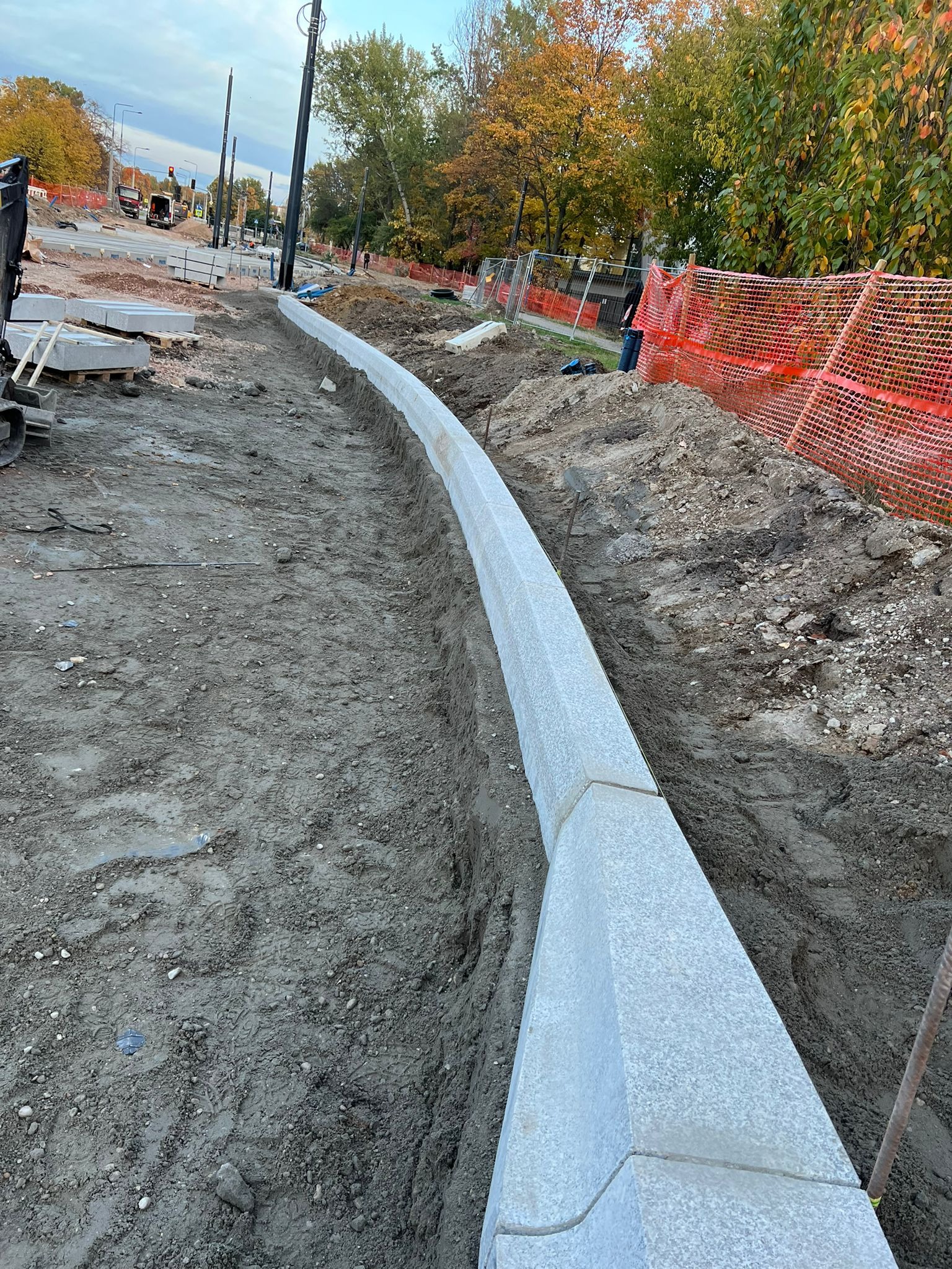 Montaż krawężnika z kostki granitowej na przygotowanym podłożu. Widoczne palety z kostką i maszyny budowlane w tle, sugerujące etap prac drogowych.