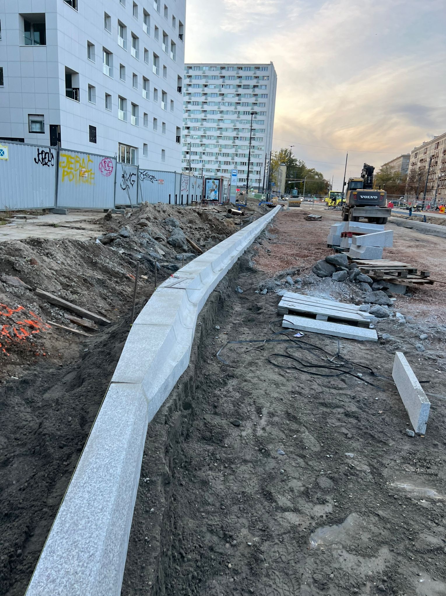 Montaż krawężników z jasnego granitu na placu budowy w miejskiej przestrzeni. W tle budynki mieszkalne i koparka Volvo.