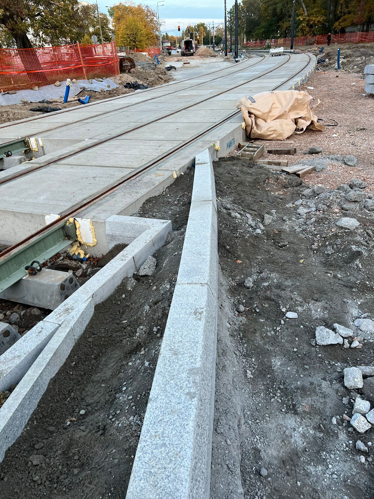 Układanie torów tramwajowych z granitowymi obrzeżami na podbudowie betonowej. Widoczne materiały budowlane i zabezpieczenia. Prace w toku, jesienna sceneria.
