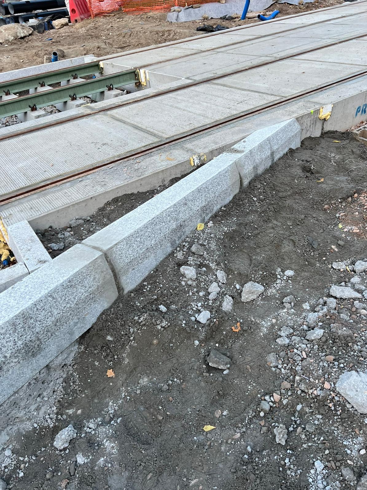 Montaż krawężnika granitowego wzdłuż betonowej konstrukcji torowiska tramwajowego. Widoczne elementy mocujące i podsypka piaskowa.