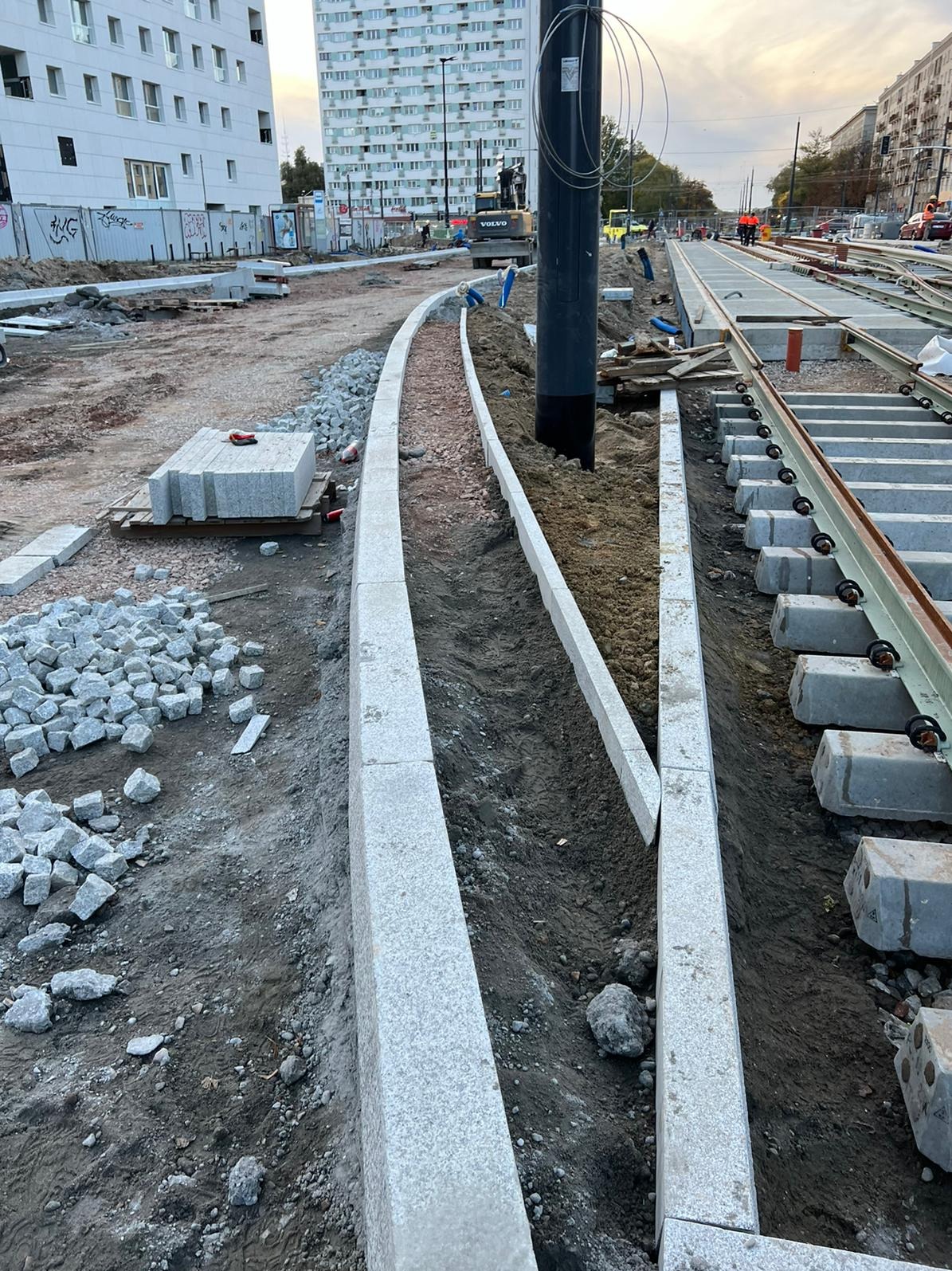 Fragment budowy torowiska tramwajowego z ułożonymi obrzeżami z kostki granitowej i przygotowaną podbudową. W tle budynki mieszkalne i sprzęt budowlany.