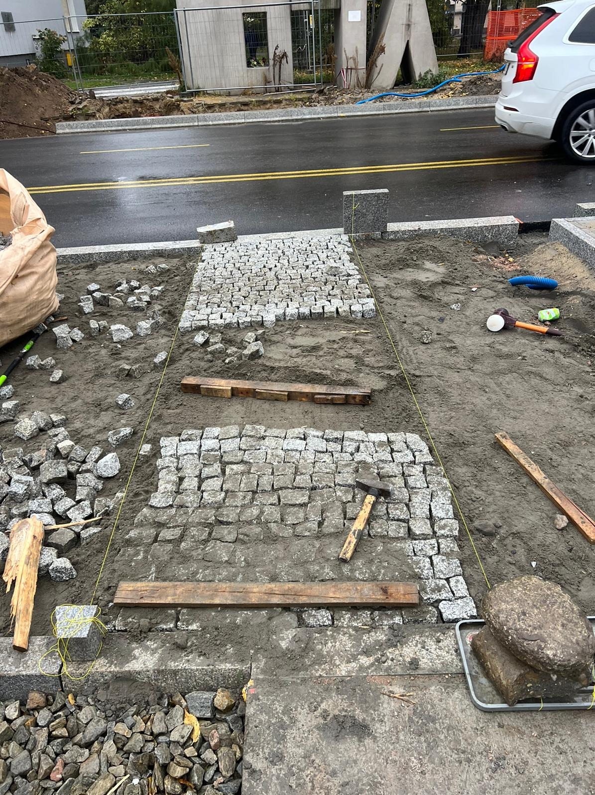 Fragment układania ścieżki z kostki granitowej, widoczne narzędzia, wytyczone linie, częściowo ułożony wzór z kostki. Praca w toku, dzień pochmurny.