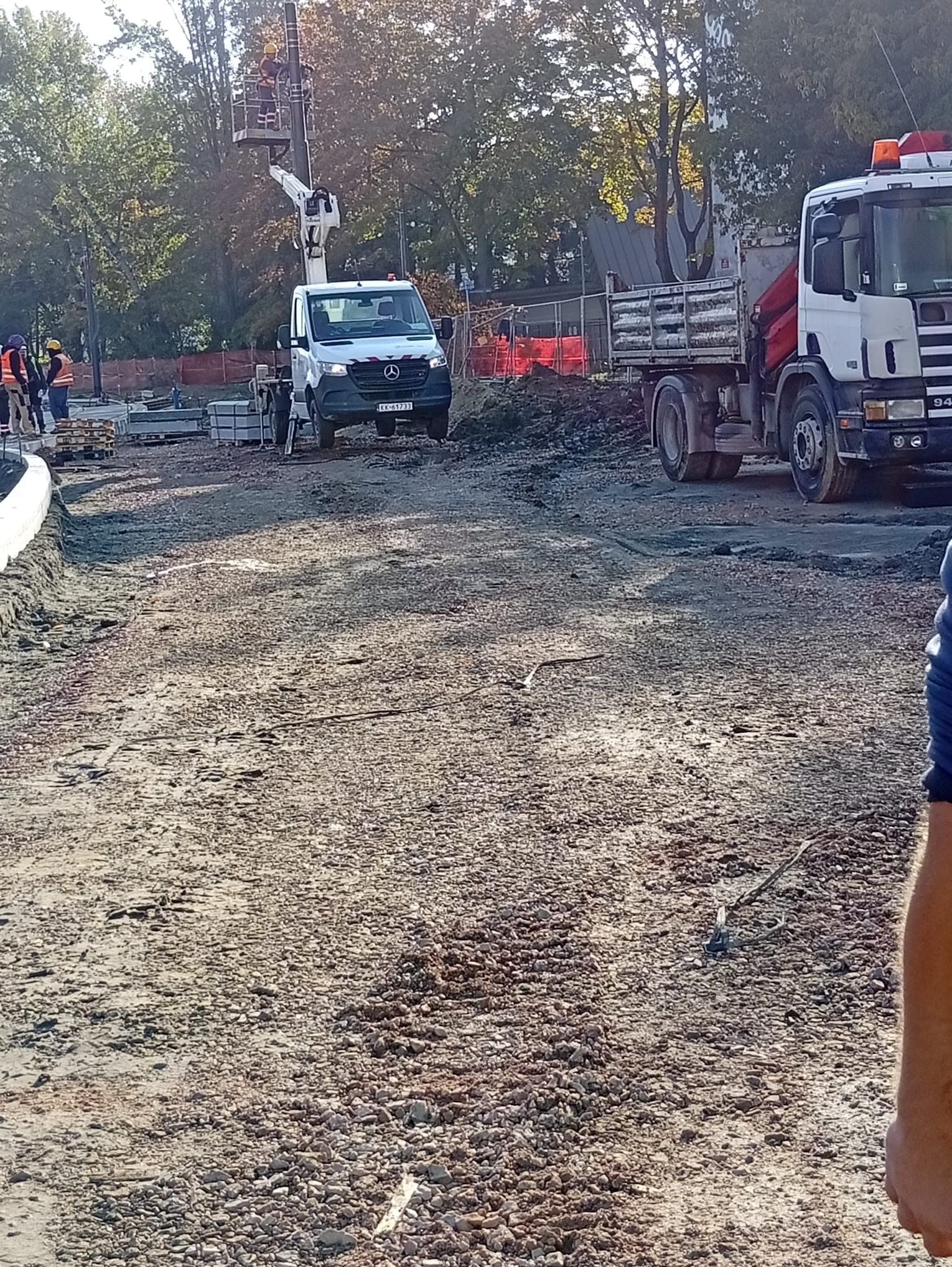 Przygotowanie terenu pod budowę: wyrównana, kamienista powierzchnia z ciężarówkami i podnośnikiem koszowym w tle. Widoczni pracownicy w odblaskowych kamizelkach.