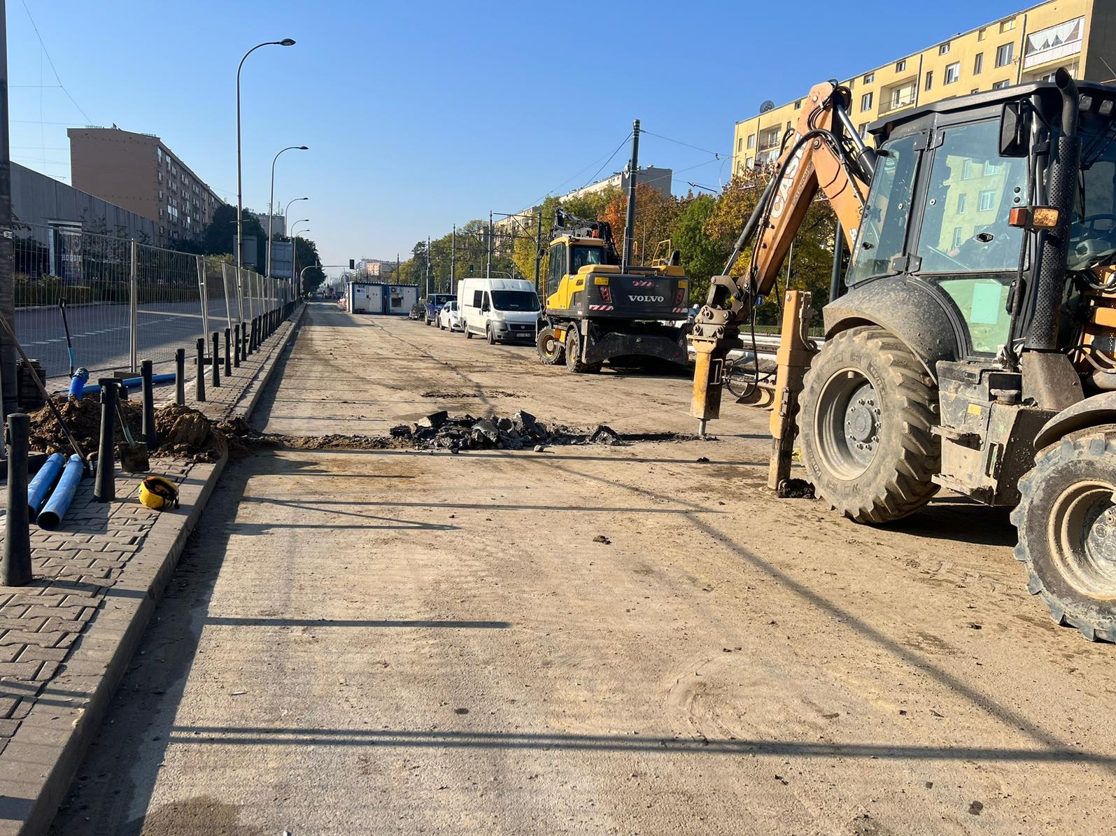 Prace ziemne na drodze miejskiej: ciężki sprzęt (koparka, młot pneumatyczny) w akcji, przygotowanie podłoża, widoczne rury i krawężnik. Słoneczny dzień.
