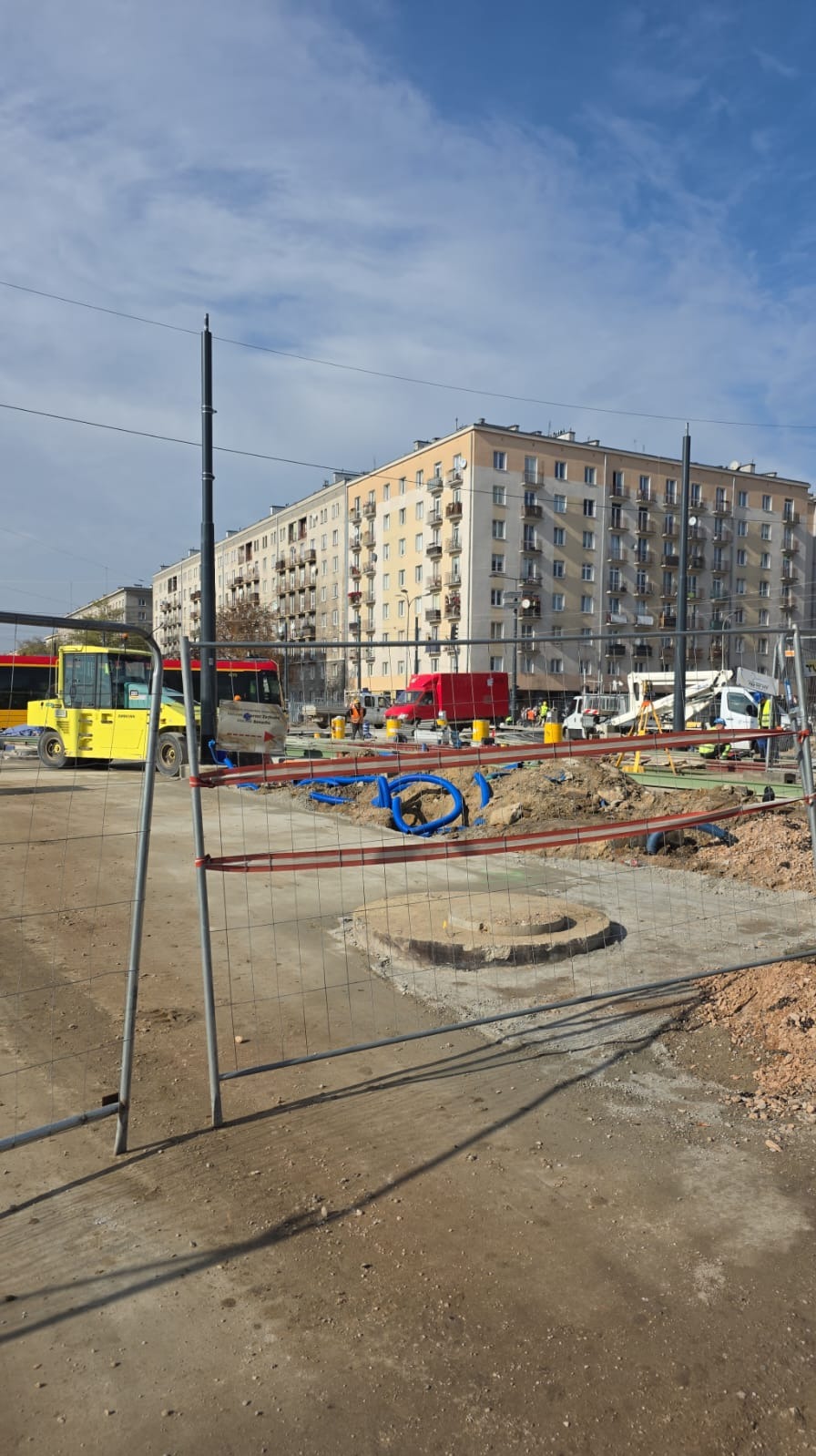 Teren budowy z wykopem, widoczne niebieskie rury, maszyny budowlane i budynek mieszkalny w tle, zabezpieczony metalowym ogrodzeniem tymczasowym.