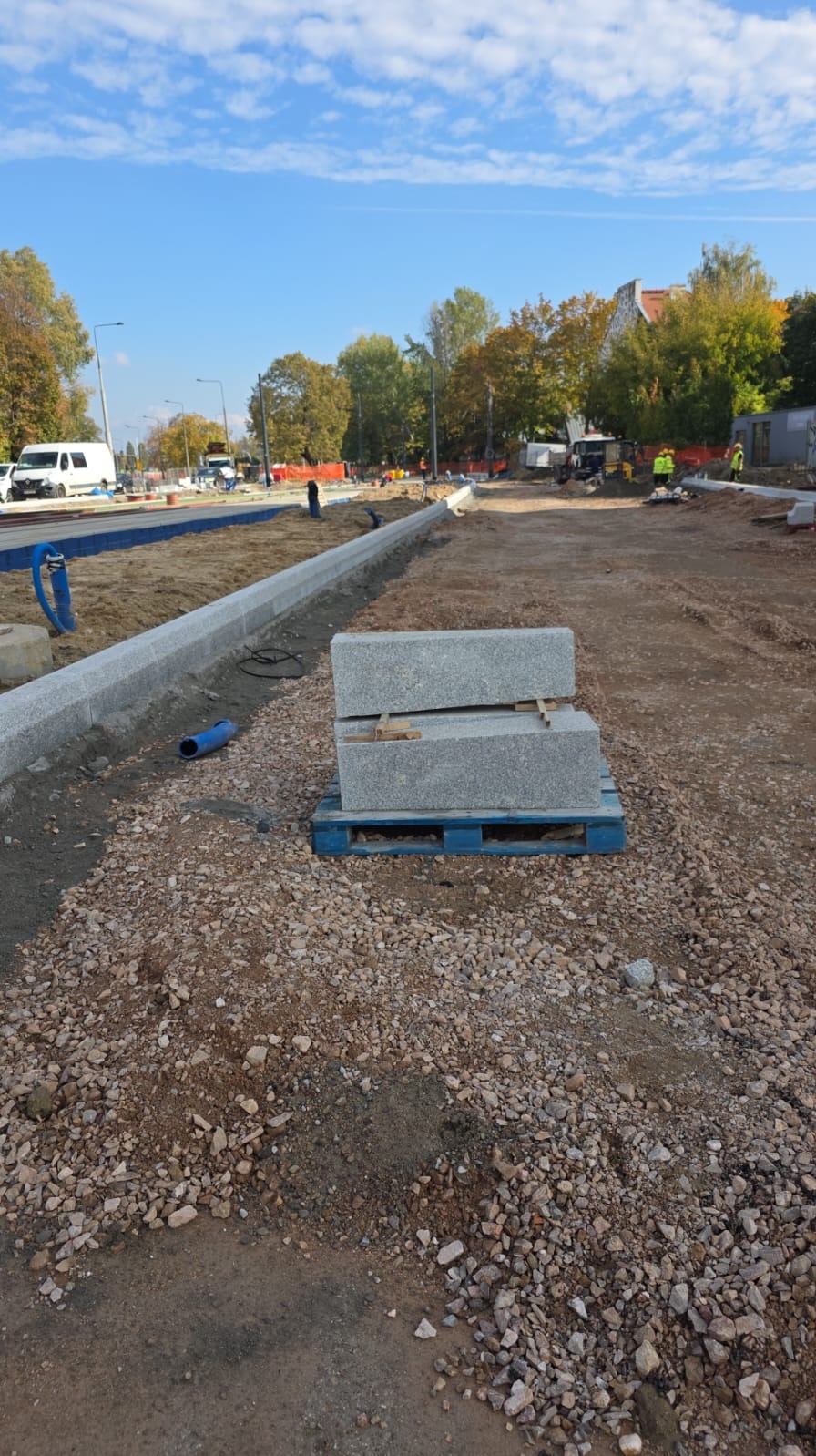 Układanie obrzeży chodnika z kostki granitowej na placu budowy. Ułożone bloki na palecie, w tle ekipa pracowników i sprzęt budowlany, błękitne niebo.