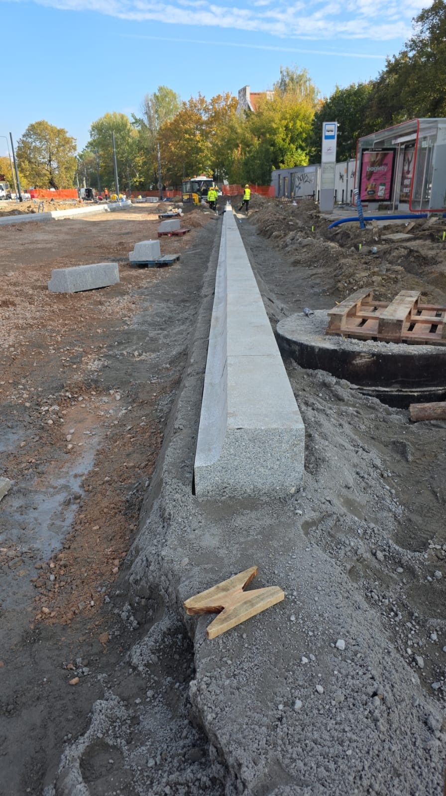 Montaż krawężnika granitowego wzdłuż drogi na przygotowanym podłożu. W tle widoczny przystanek autobusowy i drzewa.