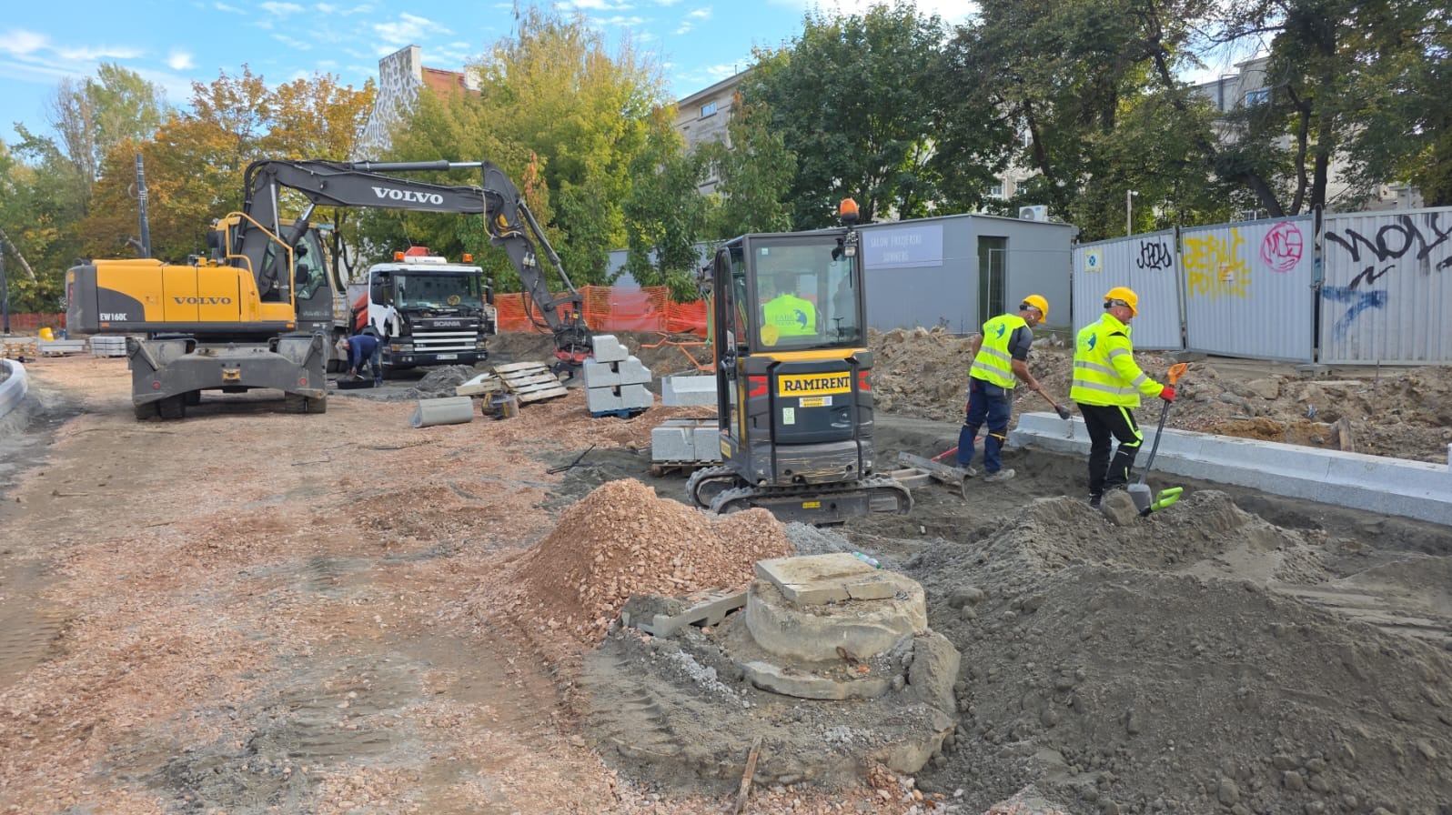 Prace ziemne przy użyciu koparek Volvo i Ramirent. Dwóch robotników w żółtych kamizelkach wyrównuje teren łopatami. Widoczne palety z kostką i fragmenty chodnika.