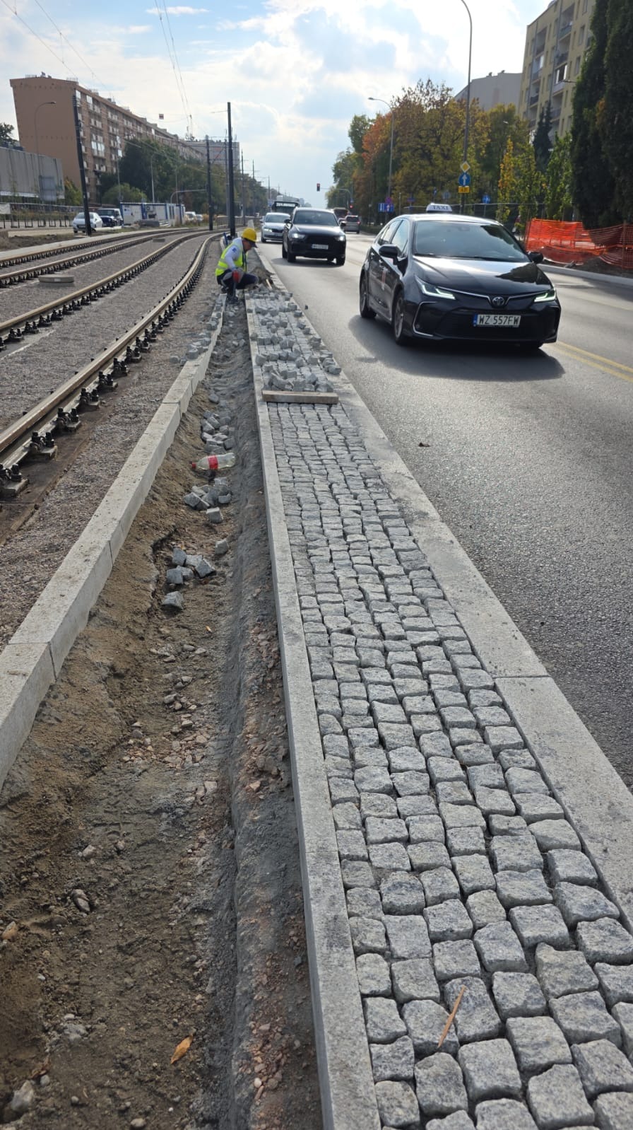 Układanie kostki granitowej przy torach tramwajowych. Widoczny fragment ułożonej nawierzchni i pracownik w żółtym kasku podczas pracy.