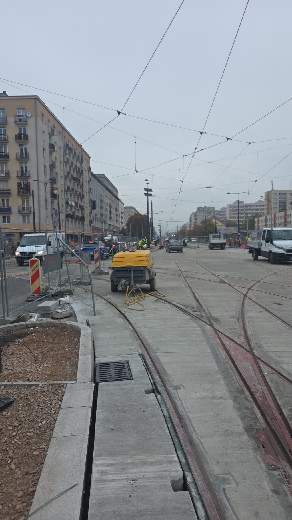 Ulica w trakcie remontu z widocznymi torami tramwajowymi, żółtym agregatem i robotnikami w oddali. W tle budynki miejskie i pochmurne niebo.
