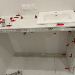 Norbert Stępień - Biała umywalka osadzona na blacie z jasnych płytek, zabezpieczonych czerwonymi klipsami. W tle białe ściany i podłoga w trakcie prac wykończeniowych w łazience.