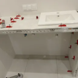 Biała umywalka osadzona na blacie z jasnych płytek, zabezpieczonych czerwonymi klipsami. W tle białe ściany i podłoga w trakcie prac wykończeniowych w łazience.